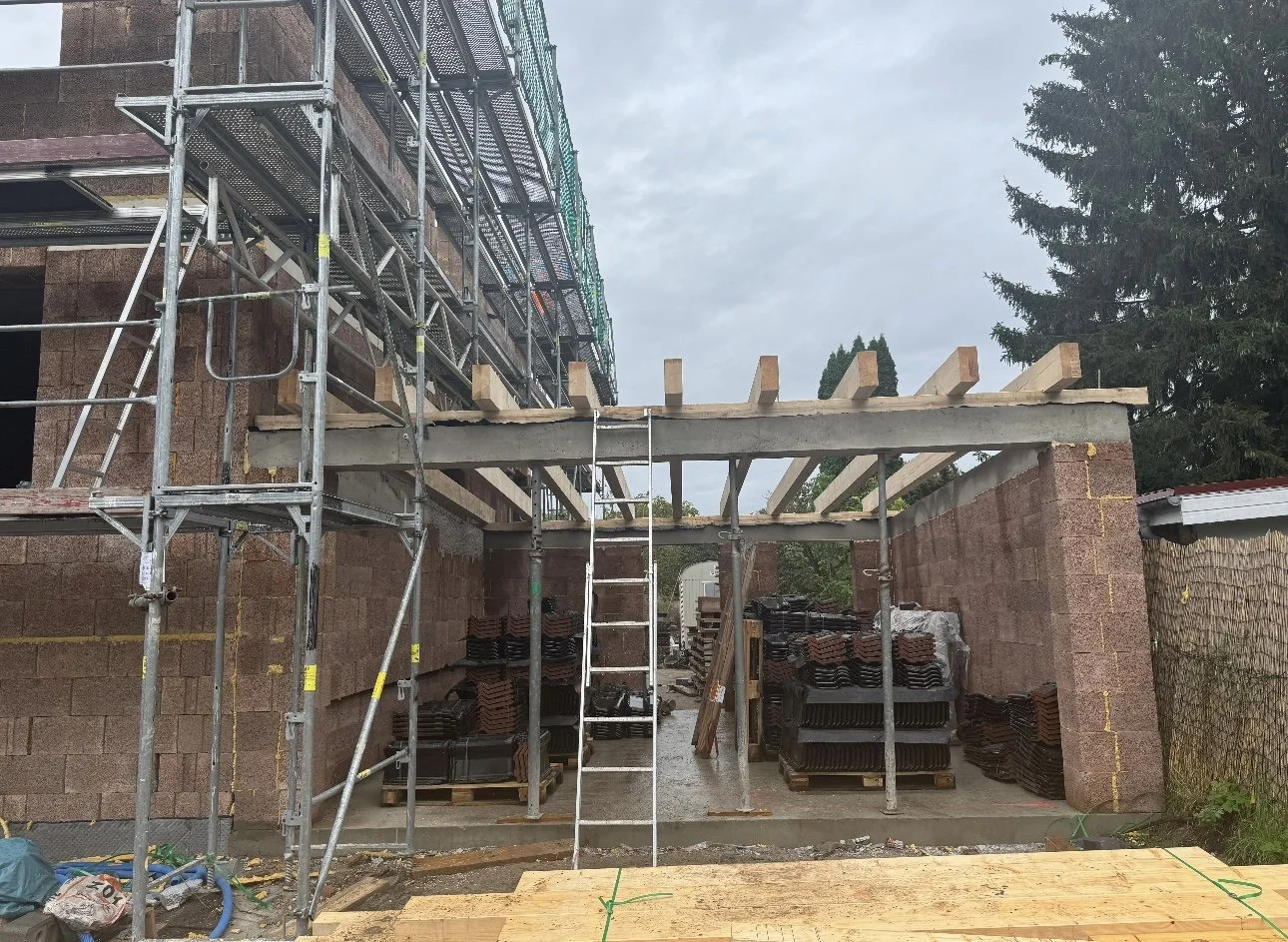 Baustelle mit Gerüsten, Holzpfosten und Baumaterialien wie Dachziegeln und Holzplatten, während das Gebäude errichtet wird.