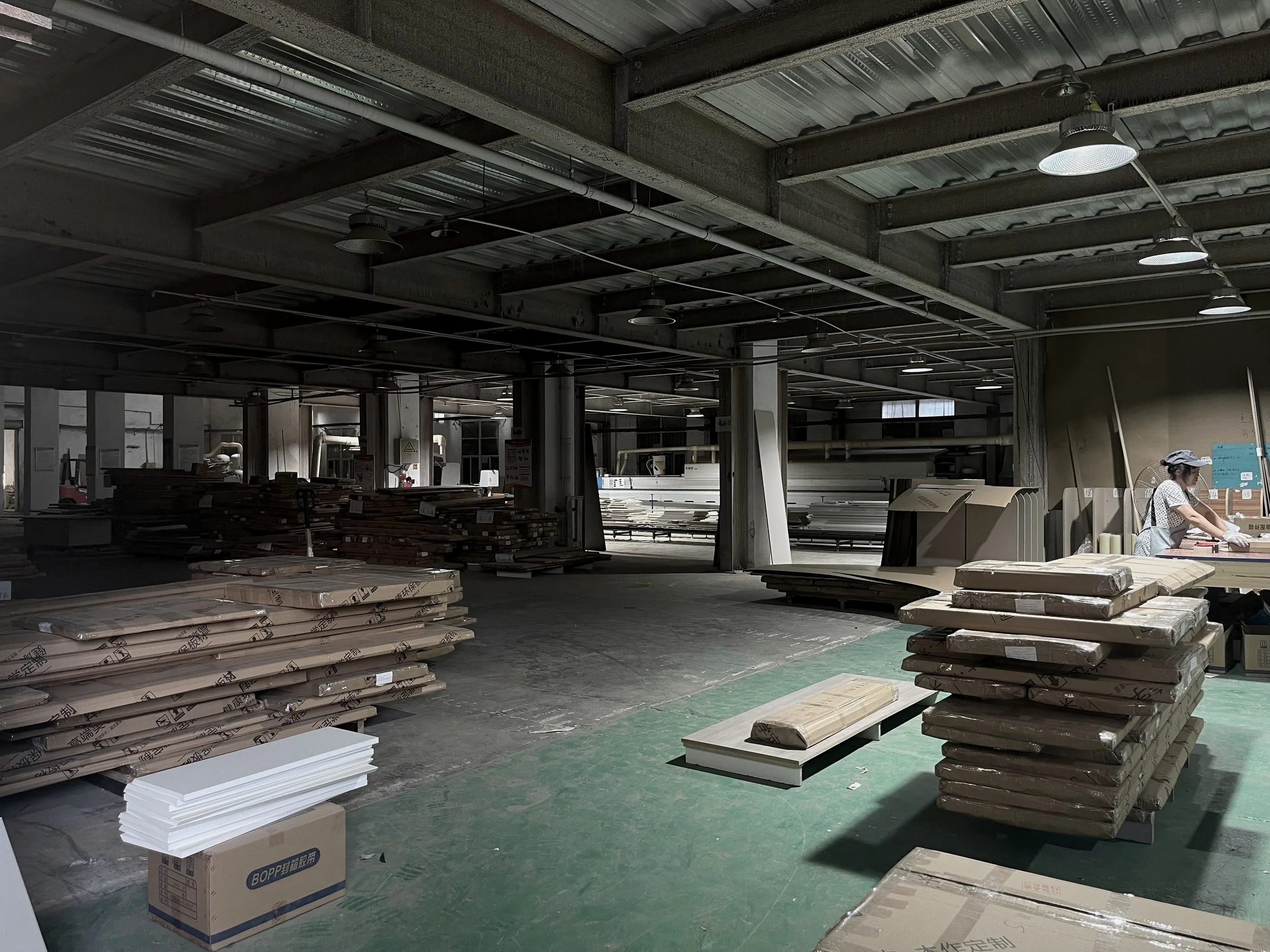 Innenraum eines Fabrik- oder Lagerhallen mit gestapelten Bau- und Möbelmaterialien, eine Frau arbeitet an einem Tisch, die Beleuchtung ist dezent, viele Materialien und Werkzeuge sind sichtbar.