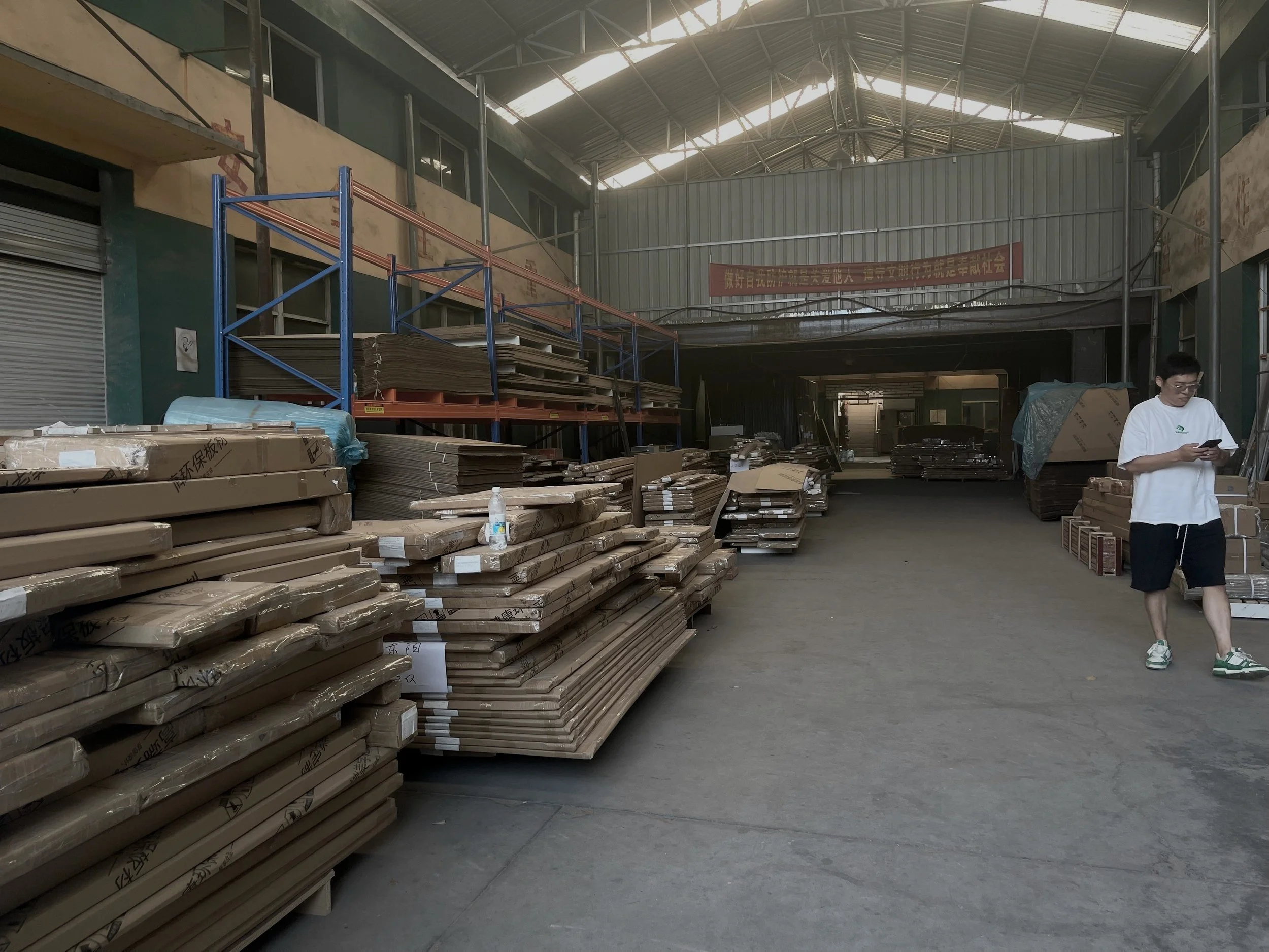 Innenansicht eines Lagerhauses mit gestapeltem Holz und Kartons, eine Person steht mit einem Smartphone in der Hand, der Raum ist groß und gut beleuchtet.