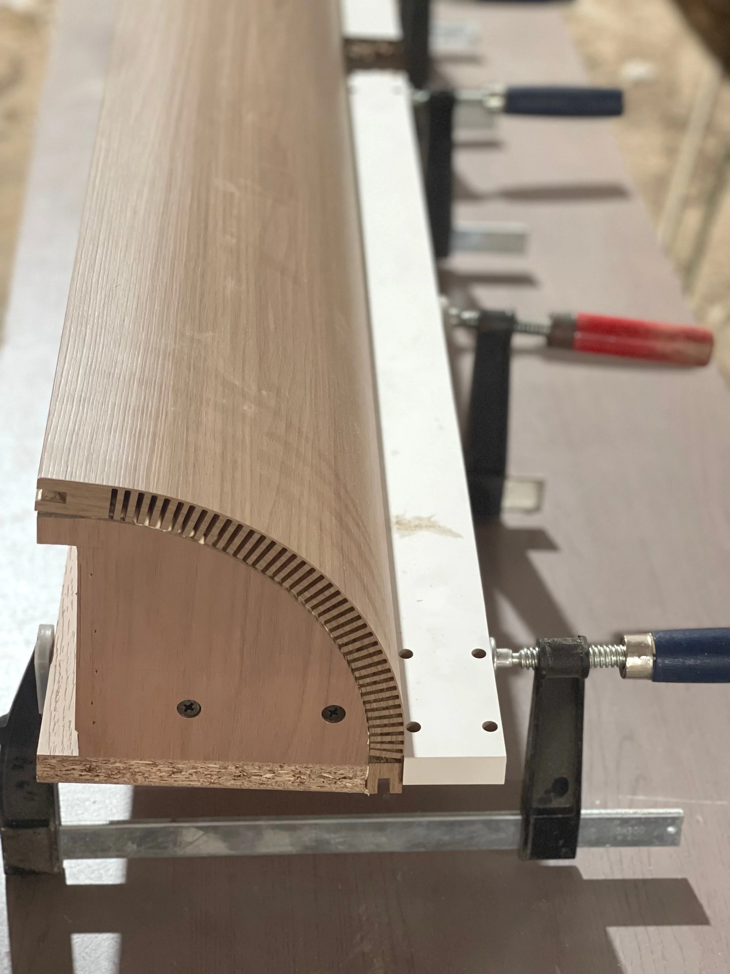 Holzstück in Form eines halbkreisförmigen Profils, das auf einer Arbeitsfläche mit Schraubzwingen befestigt ist, höchstwahrscheinlich im Rahmen einer Schreinerarbeit.