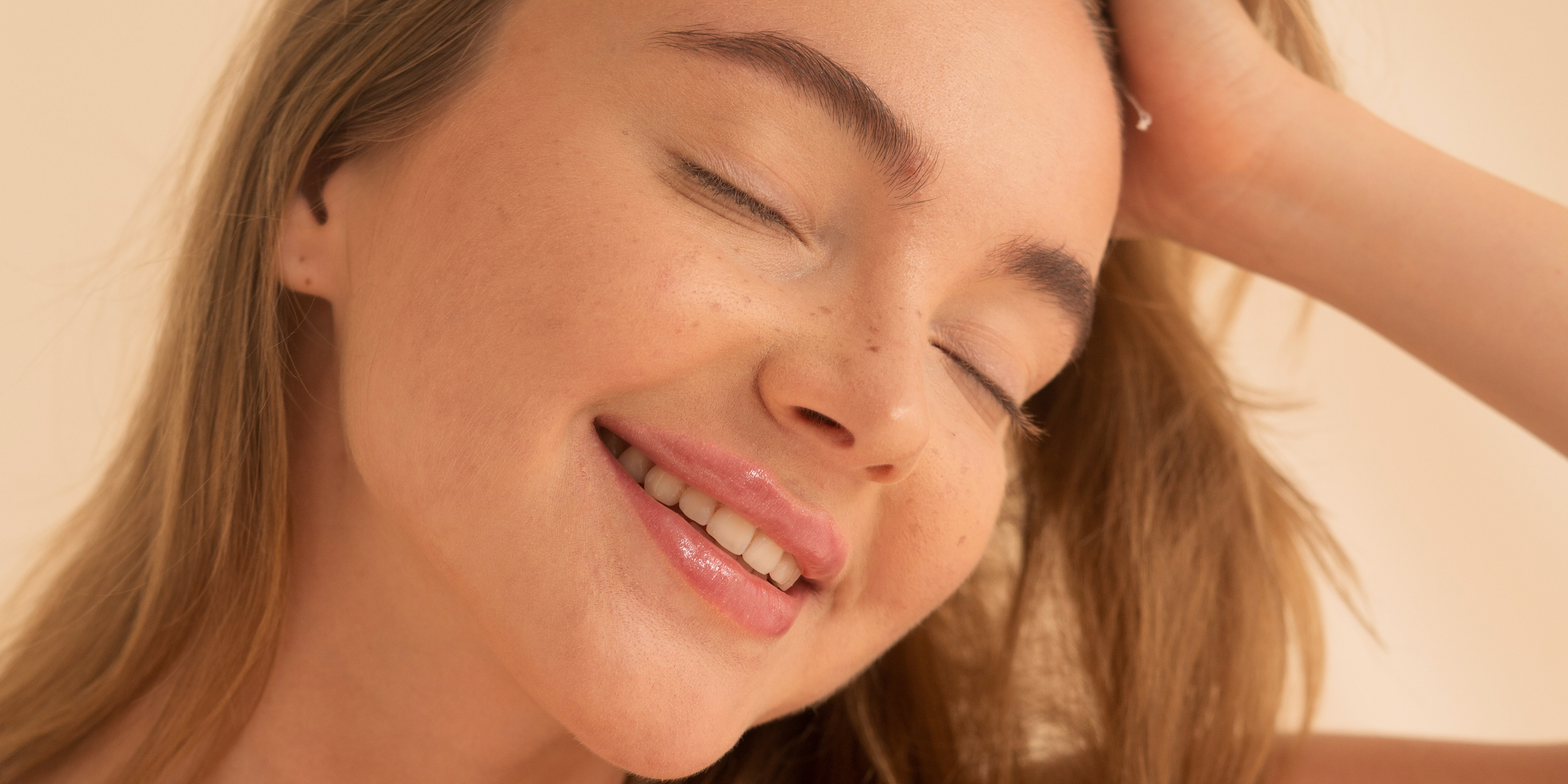 A young woman with light brown hair and freckles smiling with her eyes closed, touching her head with her arm. Botox, Fillers & Sculptra | Beauty Blueprint Scottsdale, Arizona