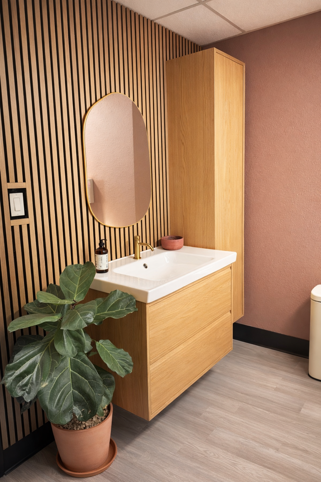 Modern bathroom with wooden wall paneling, an oval mirror with a gold frame, a white sink with a gold faucet, a green potted plant, and a small pink bowl on the sink countertop. Botox, Fillers & Sculptra | Beauty Blueprint Scottsdale, Arizona