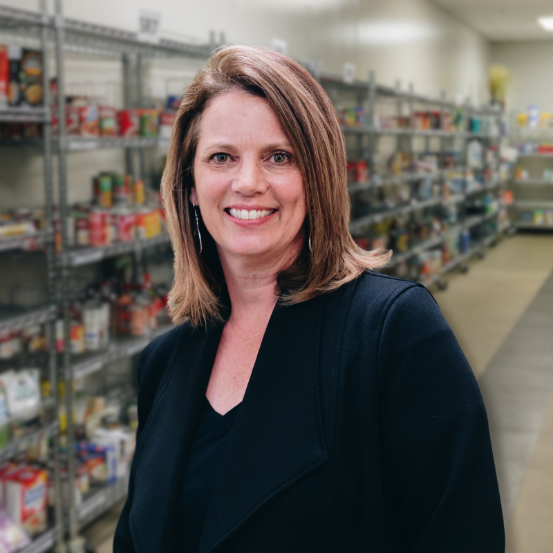 Beth is a white woman with brown hair. She is wearing a black blouse and black jacket with shelves in the background.
