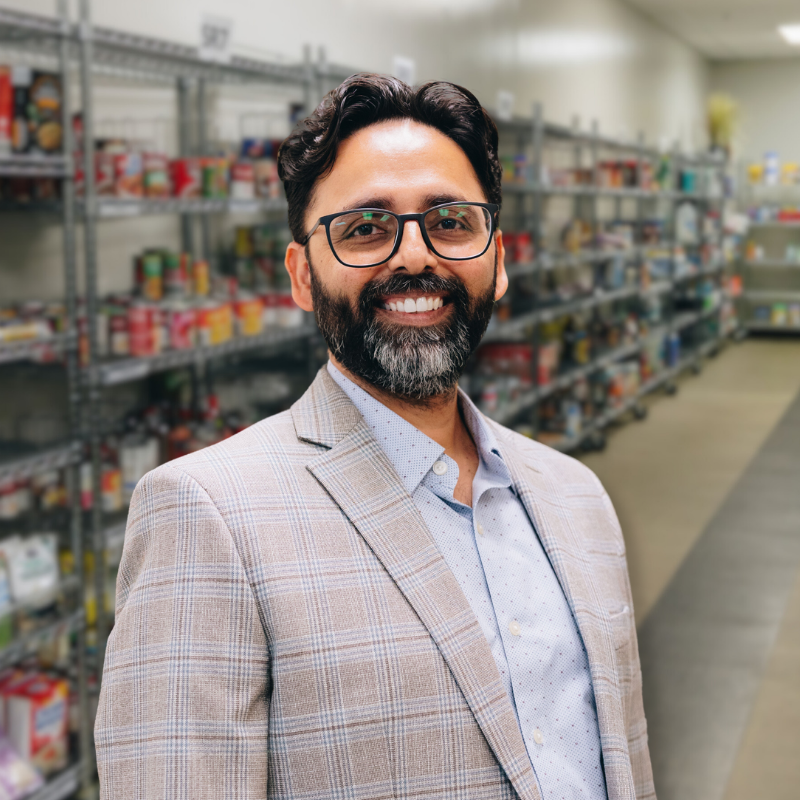 Renis is an Indian man with glasses, black hair and a short beard. He is wearing a button-up shirt with a gray suit jacket and shelves in the background.