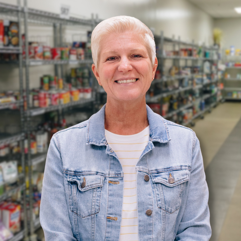 Kelli is a white woman with short, white-ish hair. She is wearing a white shirt with light gray stripes, and a denim jacket, with shelves in the background.