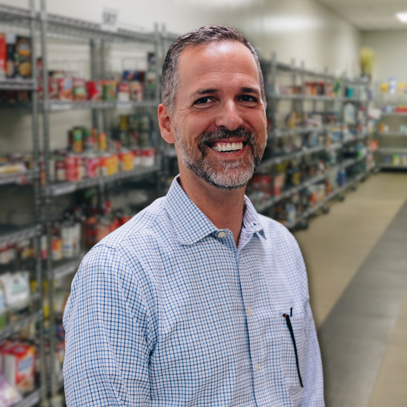 John is a white man with black and gray hair and a short beard. He is wearing a blue checkered, collared shirt with shelves in the background.