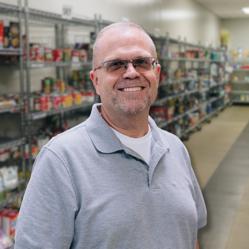 Tim is a white man with short hair and stubbly facial hair. He is wearing a gray polo with shelves in the background.