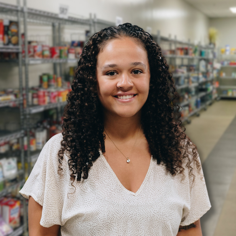 Delaney is a Black woman with long black hair. She is wearing a light pink blouse with shelves in the background.