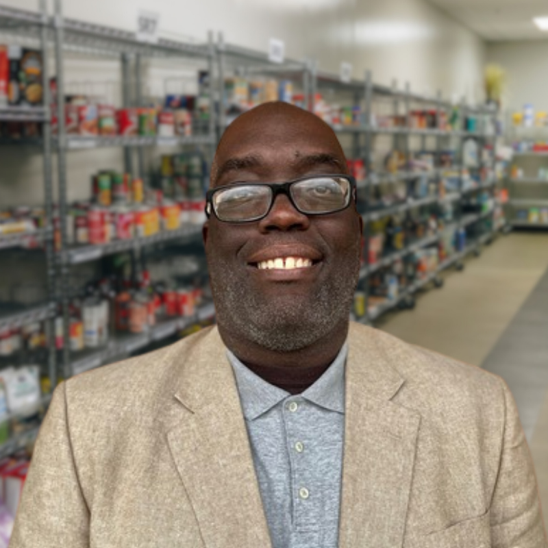 Ernest is a Black man with stubbly facial hair and glasses. He is wearing a gray collared shirt and tan jacket with shelves in the background.
