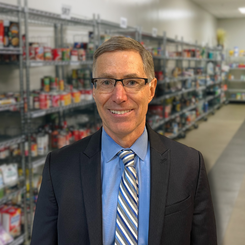 Jim is a white man with shorter, gray-ish hair and glasses. He is wearing a blue collared shirt, tie, and black suit jacket with shelves in the background.