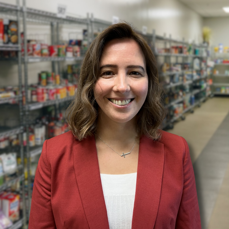 Katya is a white woman with shoulder-length brown hair. She is wearing a white blouse and maroon jacket with shelves in the background.