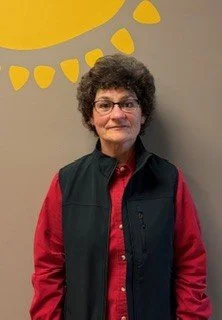 Beth Hahn is a white woman with short, brown, curly hair and glasses. She is wearing a red button-up shirt with a black vest.