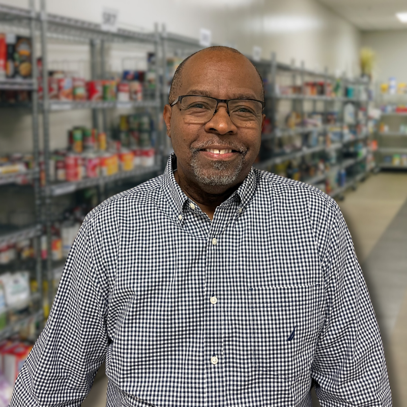 Grady is a Black man with glasses, short black hair and stubbly facial hair. He is wearing a black checkered, collared shirt with shelves in the background.