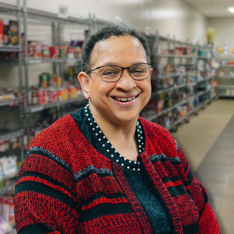 Aletha is a Black woman with short black hair and glasses. She is wearing a black blouse and red and black sweater with shelves in the background.