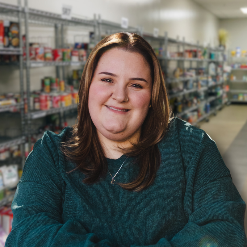 Katlyn is a white woman with long brown hair. She is wearing a dark green sweater with shelves in the background.