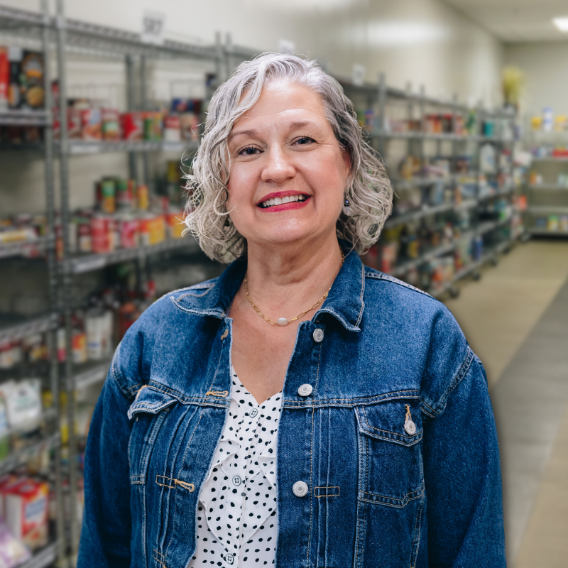 Tracy is a white woman with shorter, gray hair. She is wearing a polka dot blouse and denim jacket with shelves in the background.