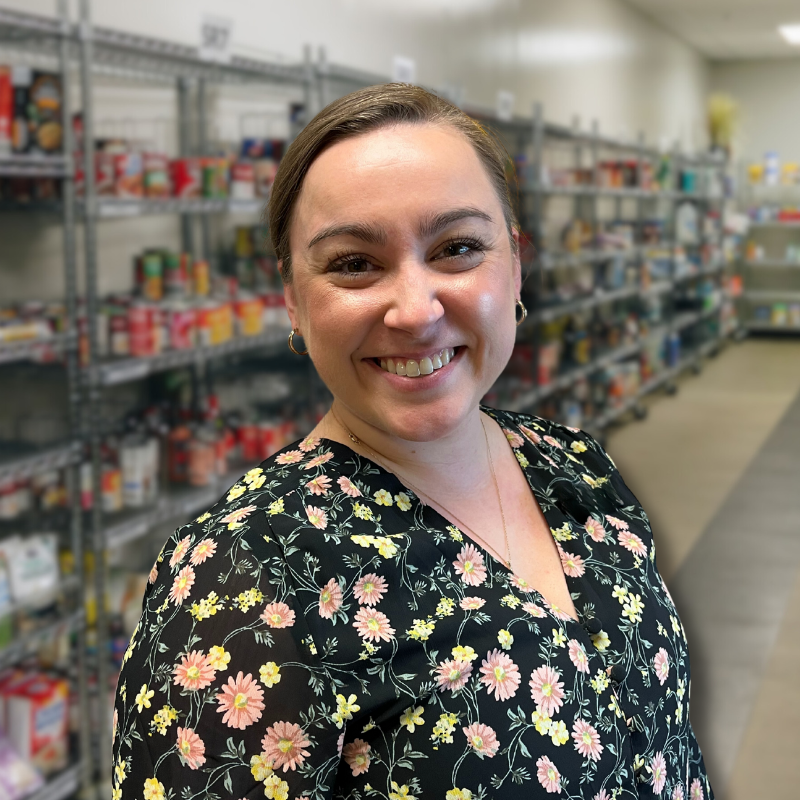 Abby is a white woman with long brown hair in a bun. She is wearing a floral blouse with shelves in the background.
