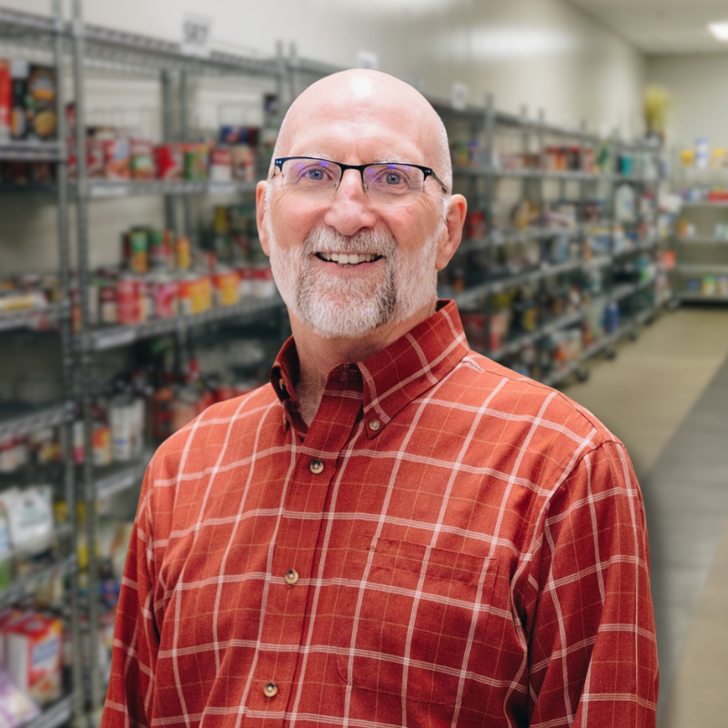 David is a white man with glasses and a short beard. He is wearing a red checkered, collared shirt with shelves in the background.
