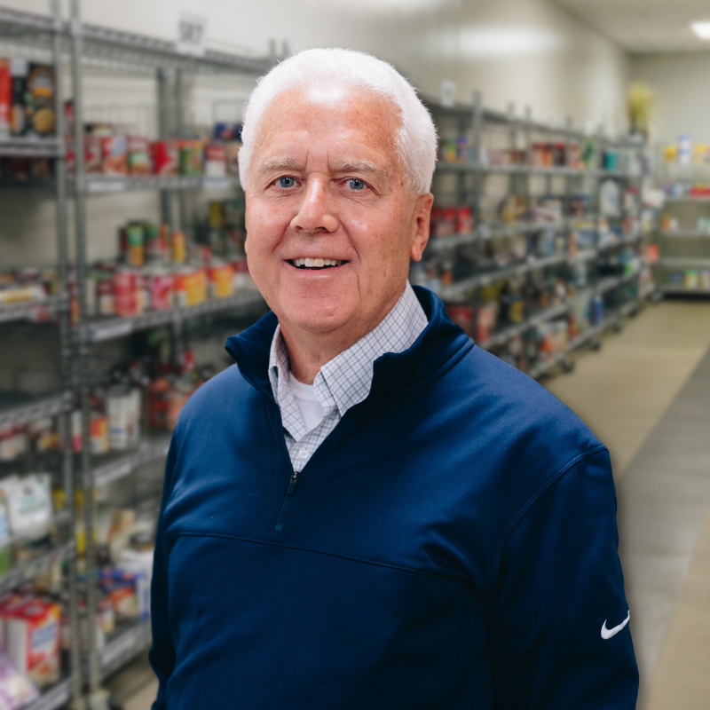 Glenn is a white man with white hair. He is wearing a collared shirt with a blue quarter zip and shelves in the background.