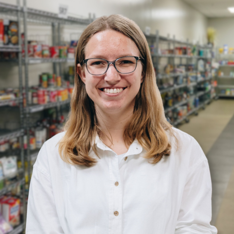 Amber is a white woman with glasses and shoulder length blonde hair. She is wearing a white, collared, button up shirt with shelves in the background.