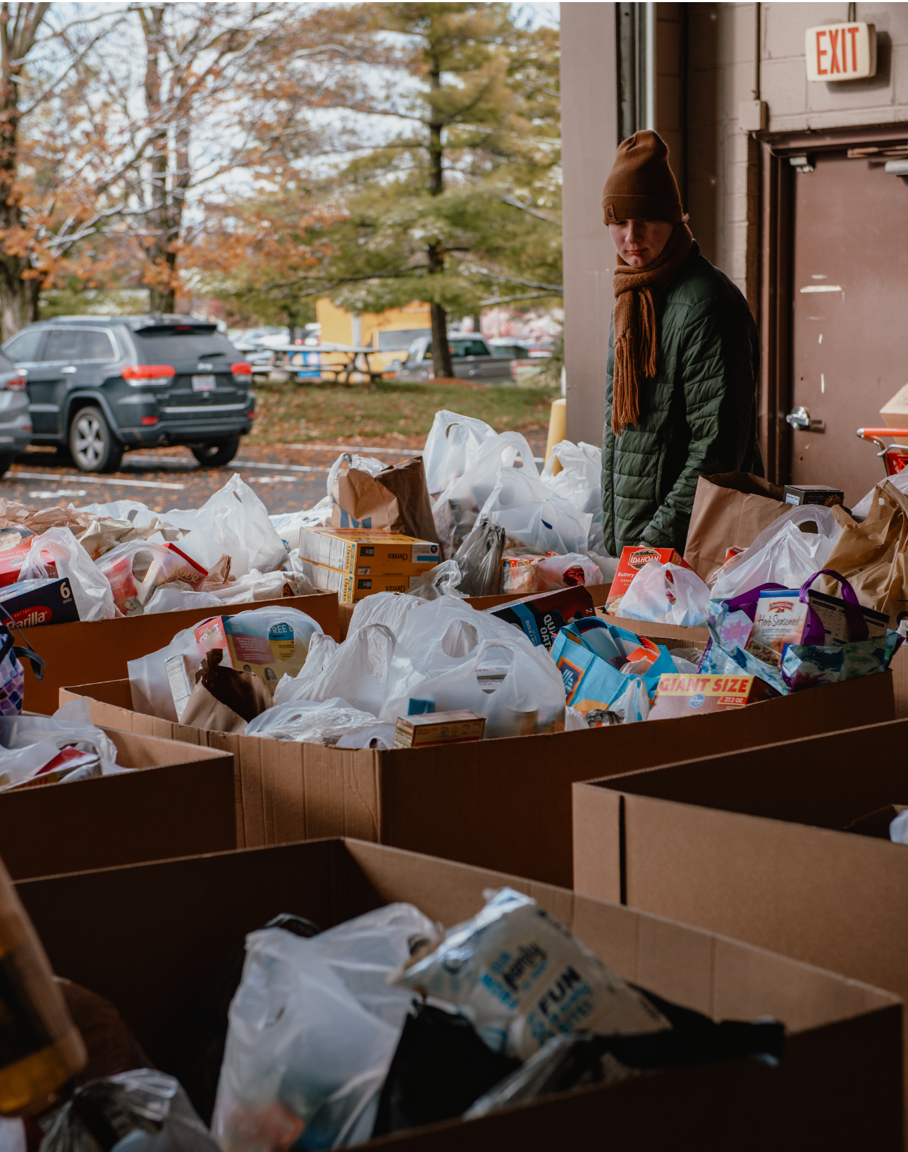 A person in a black jacket with a brown hat and scarf is standing amongst boxes of food donations in the warehouse.