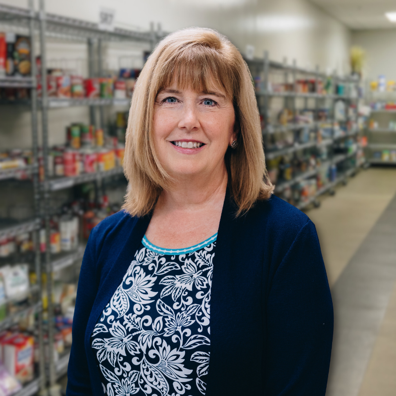 Joyce is a white woman with shoulder-length blonde hair. She is wearing a floral blouse and navy sweater with shelves in the background.