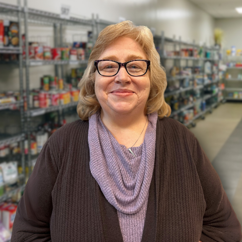 Rebecca is a white woman with glasses and shoulder length blonde hair. She is wearing a dark brown cardigan over a purple sweater with shelves in the background.