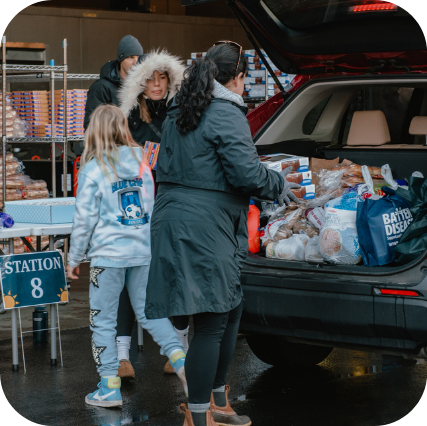 3 volunteers of varying ages, bundled for the cold, are packing food into a client's trunk for Thanksgiving Blessing.