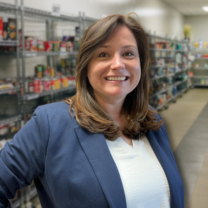 Sara is a white woman with shoulder length brown hair. She is wearing a white blouse with a navy jacket and shelves in the background.