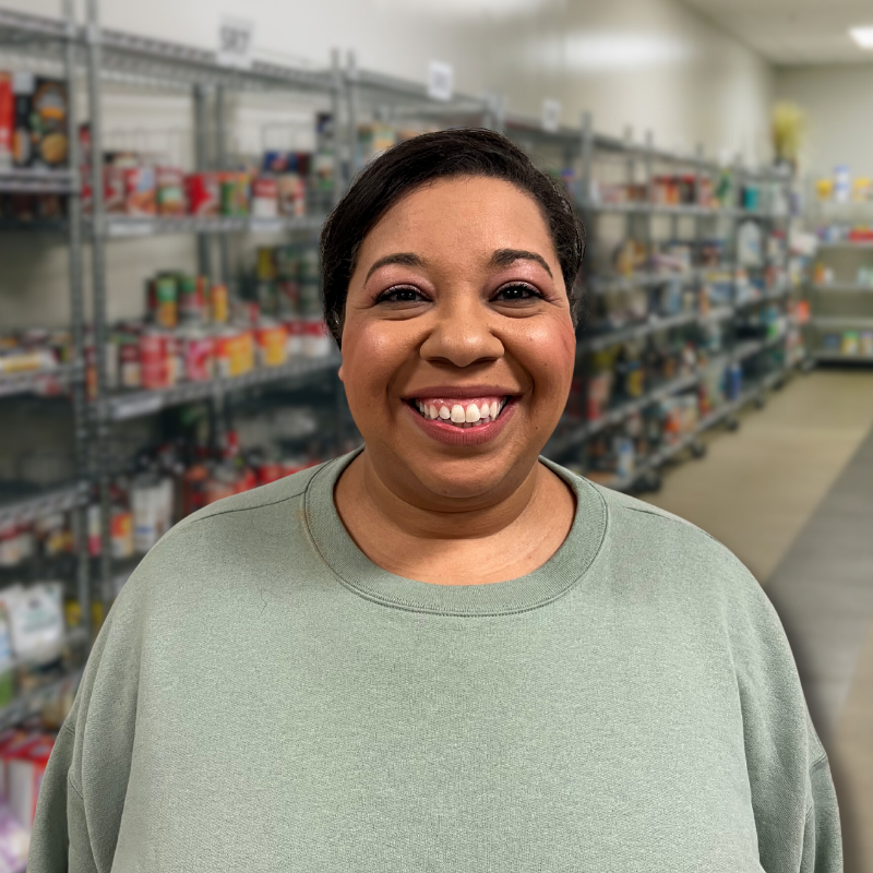Sarah is a Black woman with black hair in a bun. She is wearing a light green sweater with shelves in the background.