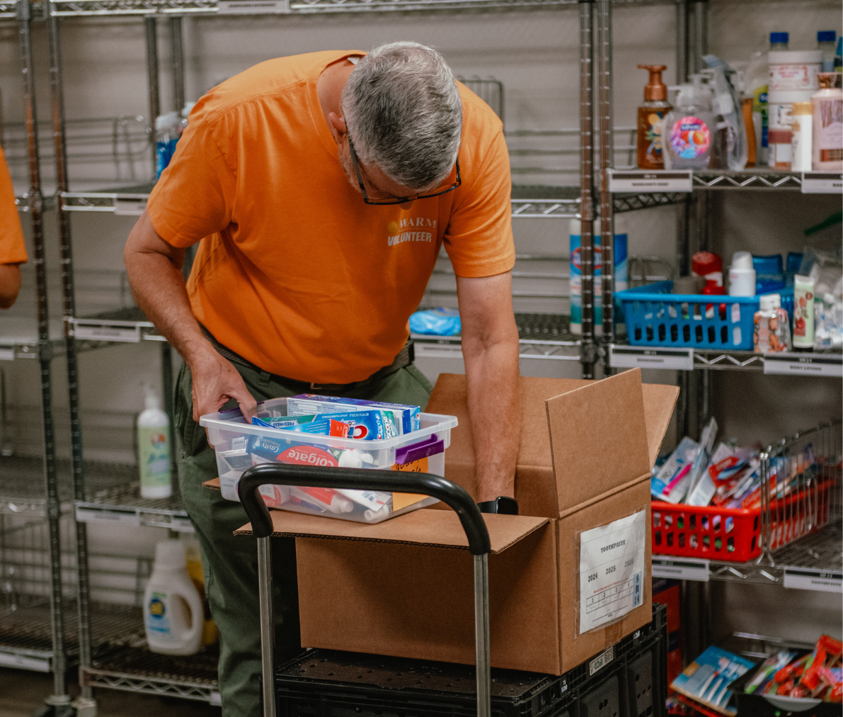 A volunteer in an orange volunteer shirt is looking in a cardboard box to stock the market shelves. He is holding a tub of toothpaste.