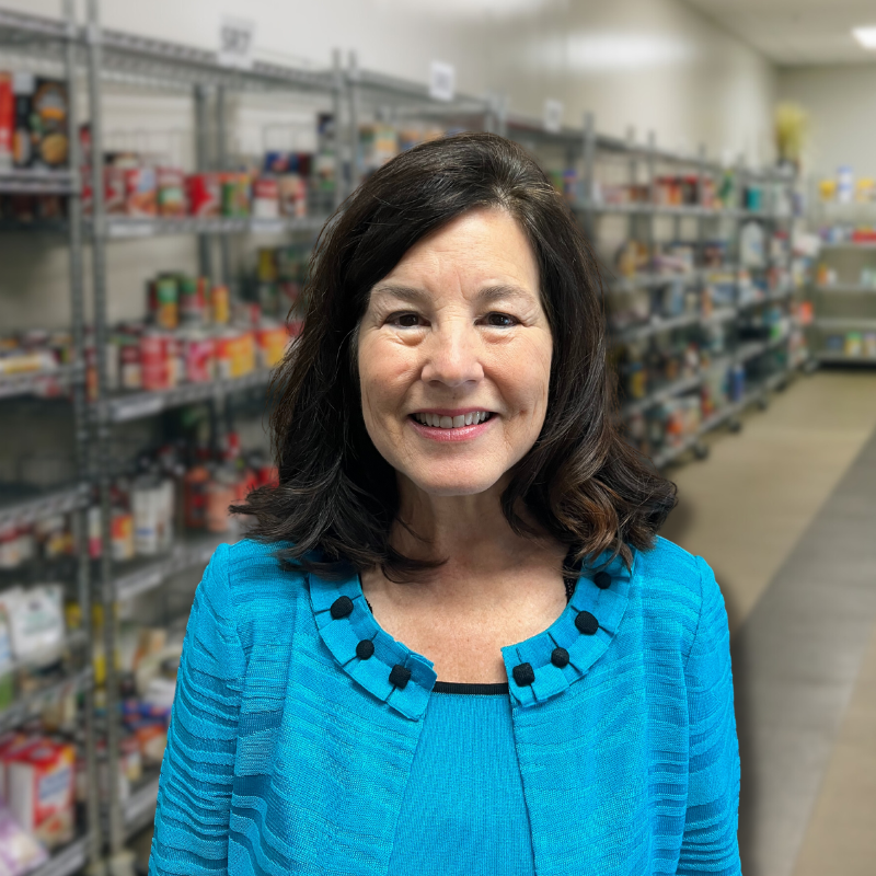 Patti is a white woman with shoulder length black hair. She is wearing a light blue blouse with shelves in the background.