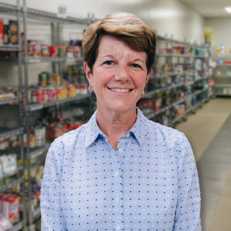 Jami is a white woman with short, brown hair. She is wearing a blue, polka dot blouse with shelves in the background.