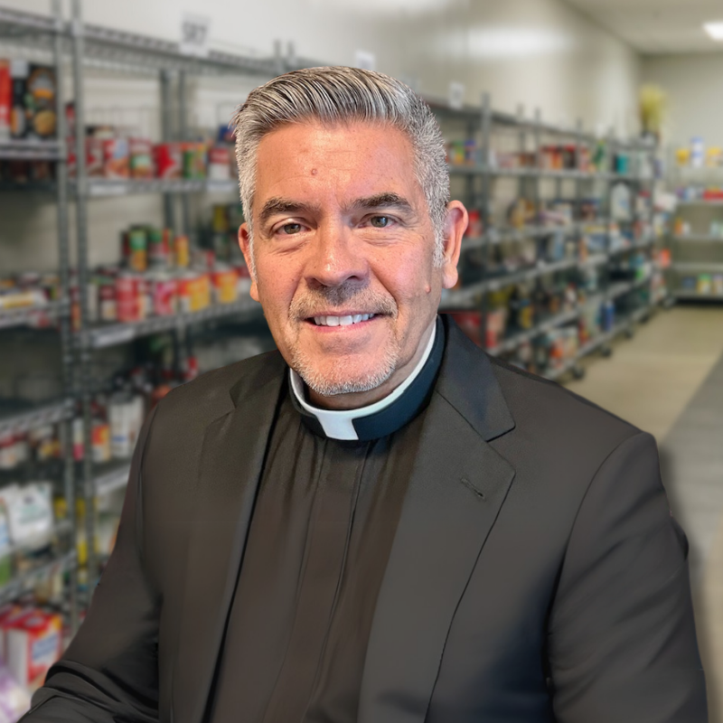 David is a white man with gray hair and stubbly facial hair. He is wearing a black shirt and clerical collar with a black jacket and shelves in the background.