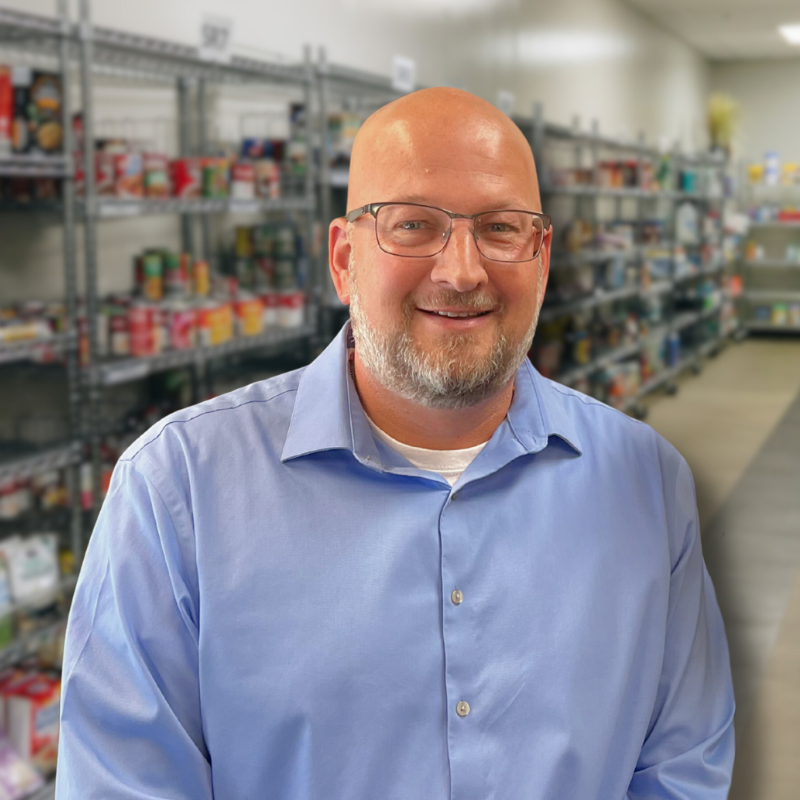Chad is a white man with glasses and a short beard. He is wearing a blue collared shirt with shelves in the background.