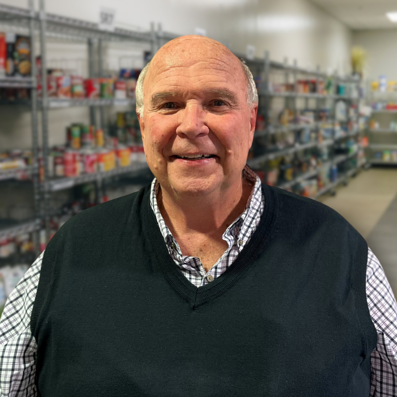 Steve is a white man with short white hair. He is wearing a blue checkered, collared shirt with a black vest, and shelves in the background.