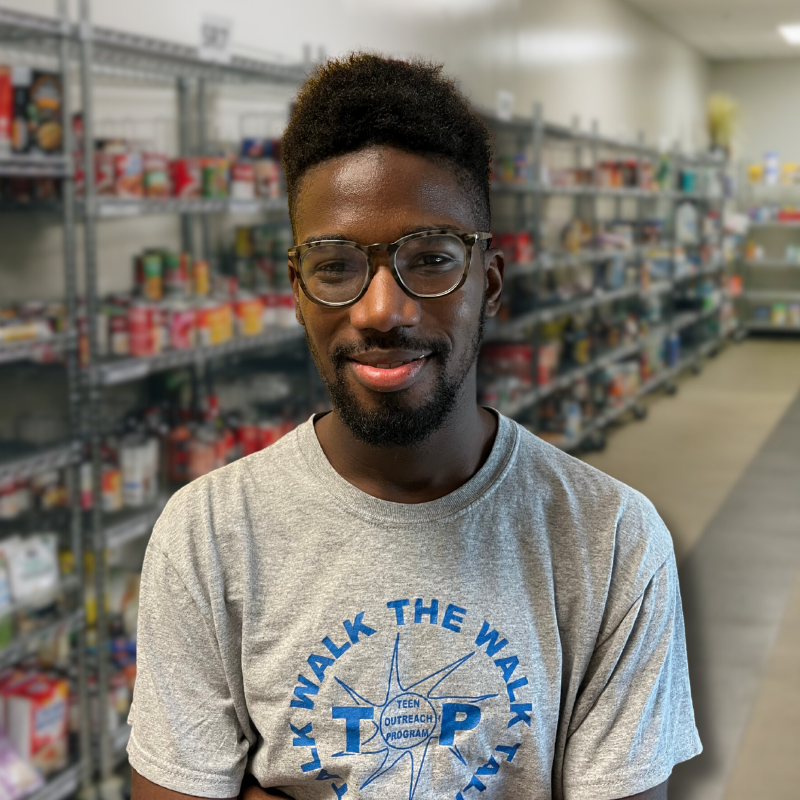 Hylan is a Black man with glasses, black hair and short facial hair. He is wearing a gray t-shirt with shelves in the background.