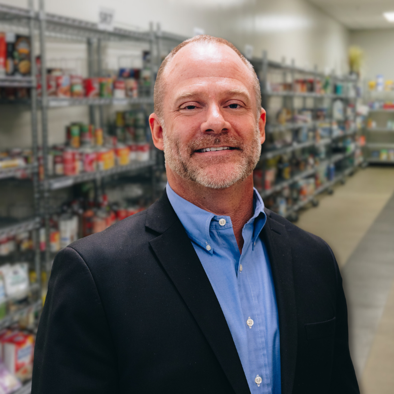 Glenn is a white man with short hair and stubbly facial hair. He is wearing a blue button up and black jacket with shelves in the background.