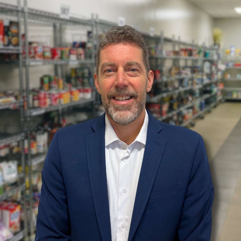 Adam is a white man with brown and gray hair with a short beard. He is wearing a white collared shirt and blue suit jacket with shelves in the background.