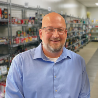 Chad Maxeiner, a white man, with glasses and stubbly facial hair. He is wearing a blue button up shirt and is in front of shelves in our choice market.