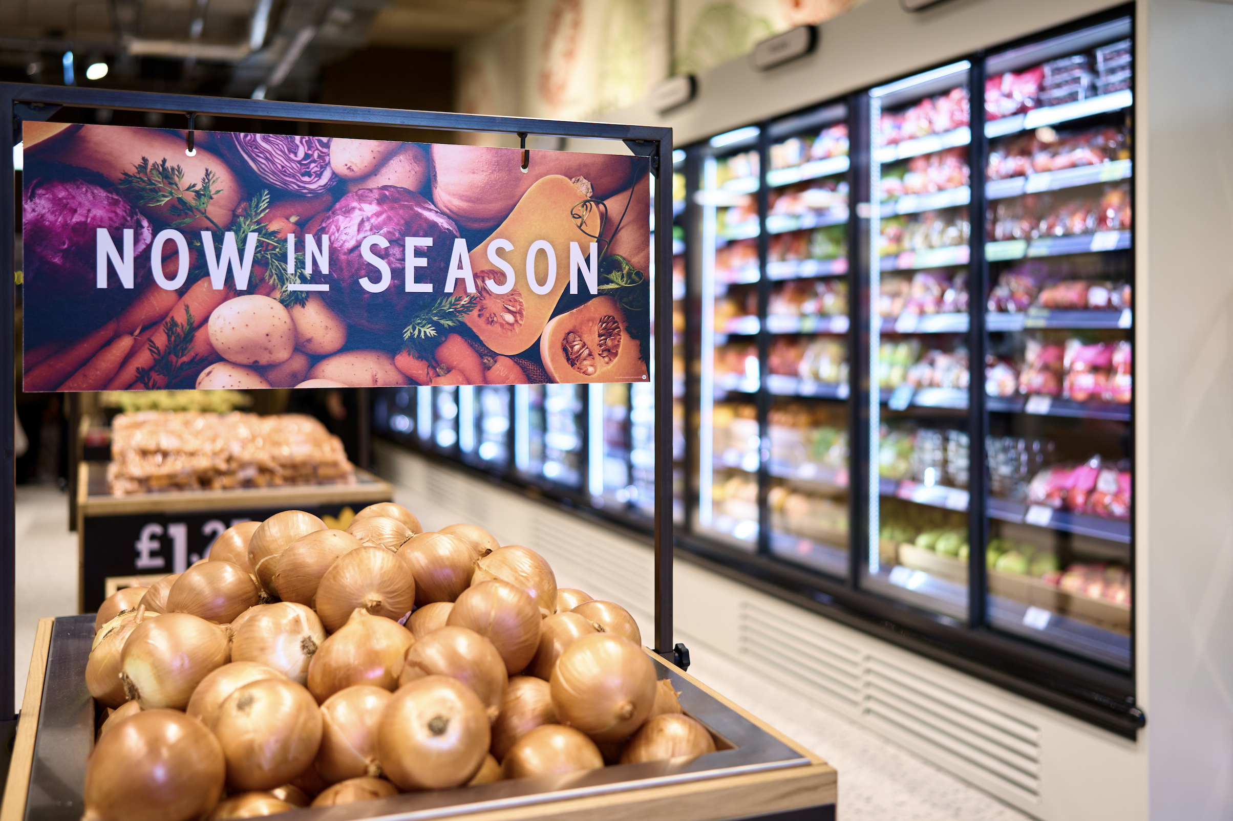 Fresh onions displayed in a produce section of a M&S store with a sign that reads 'Now in Season' and an array of cold storage units filled with produce in the background.
