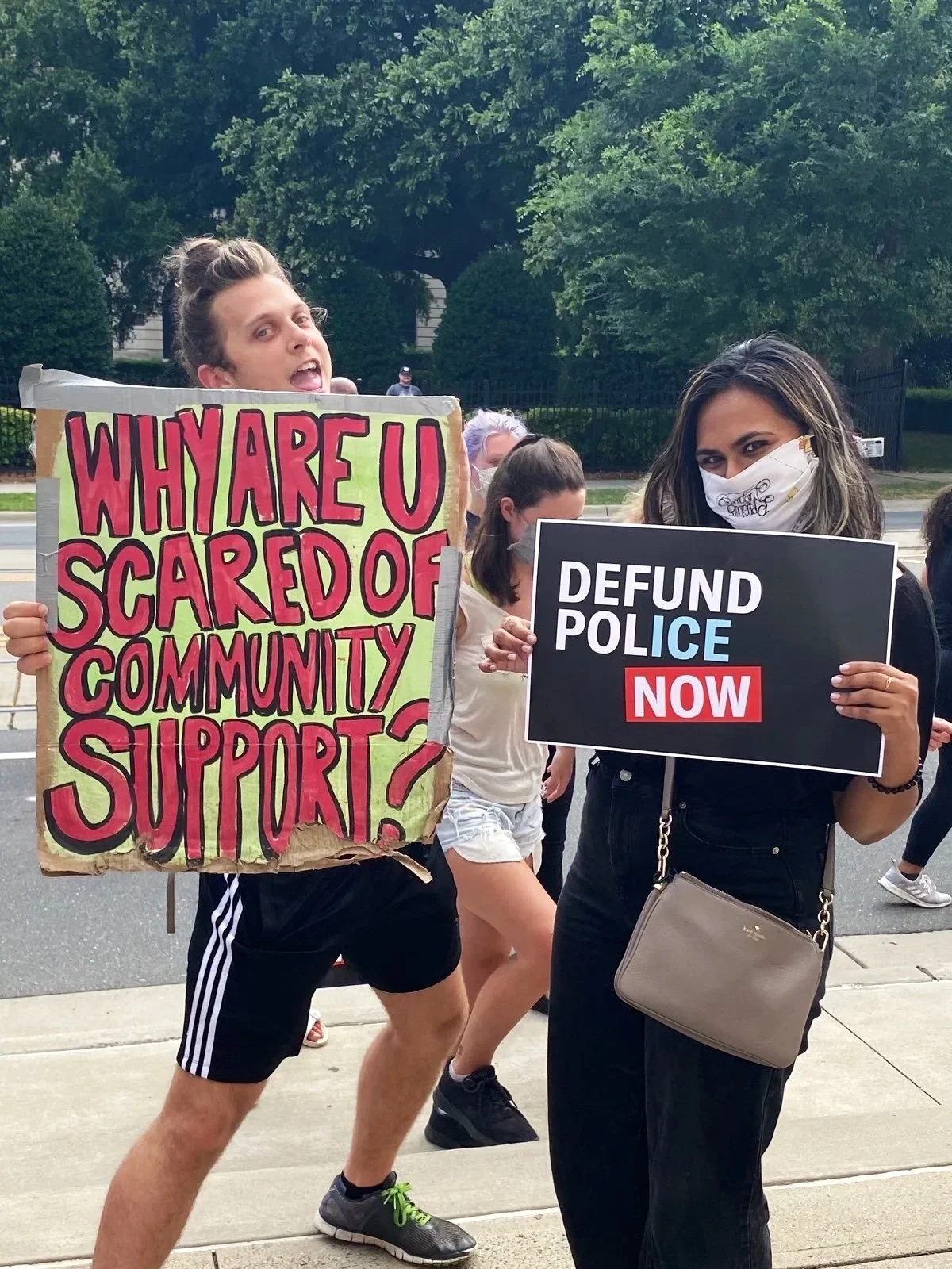 Two women at a protest holding signs addressing community support and police defunding, with other protesters in the background and trees lining the street.