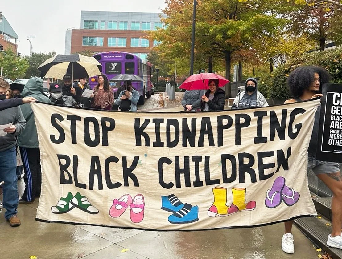 Protesters holding a banner that says "Stop Kidnapping Black Children" with illustrations of colorful sneakers, during a rainy day.