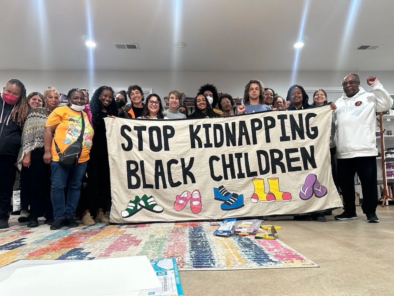 Group of people holding a banner that reads 'Stop Kidnapping Black Children,' with colorful shoe illustrations on a carpeted floor indoor setting.