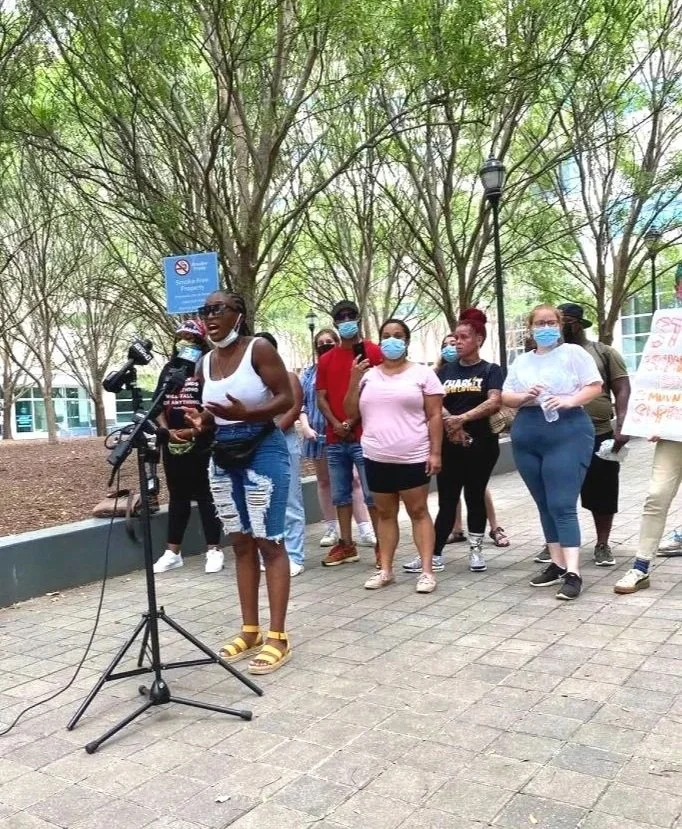 A woman speaking at a microphone during a protest or rally, surrounded by people wearing masks in an outdoor area with trees.