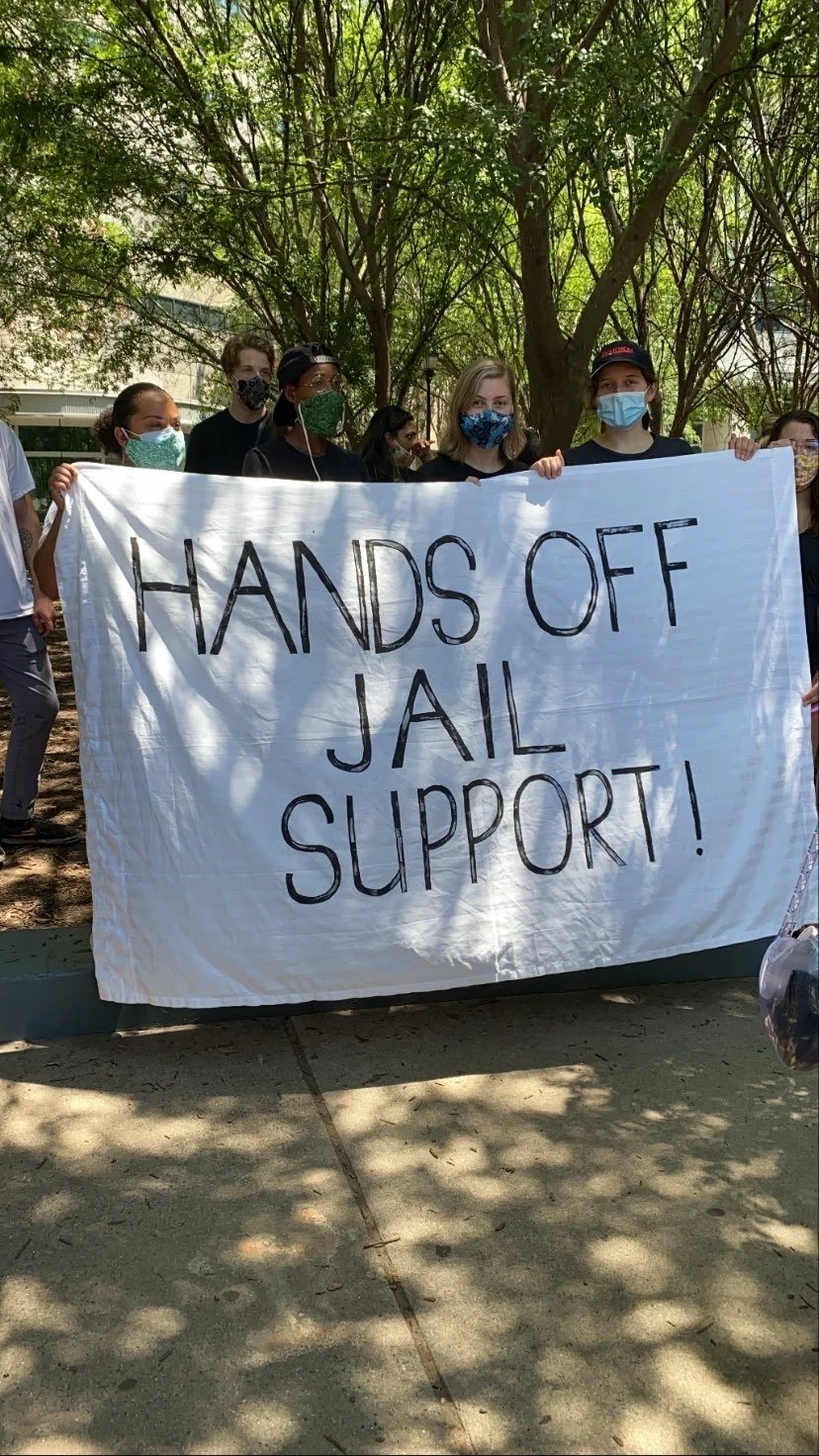 A group of people holding a white banner that reads "HANDS OFF JAIL SUPPORT!" during a protest, with trees and sunshine in the background, all wearing masks.
