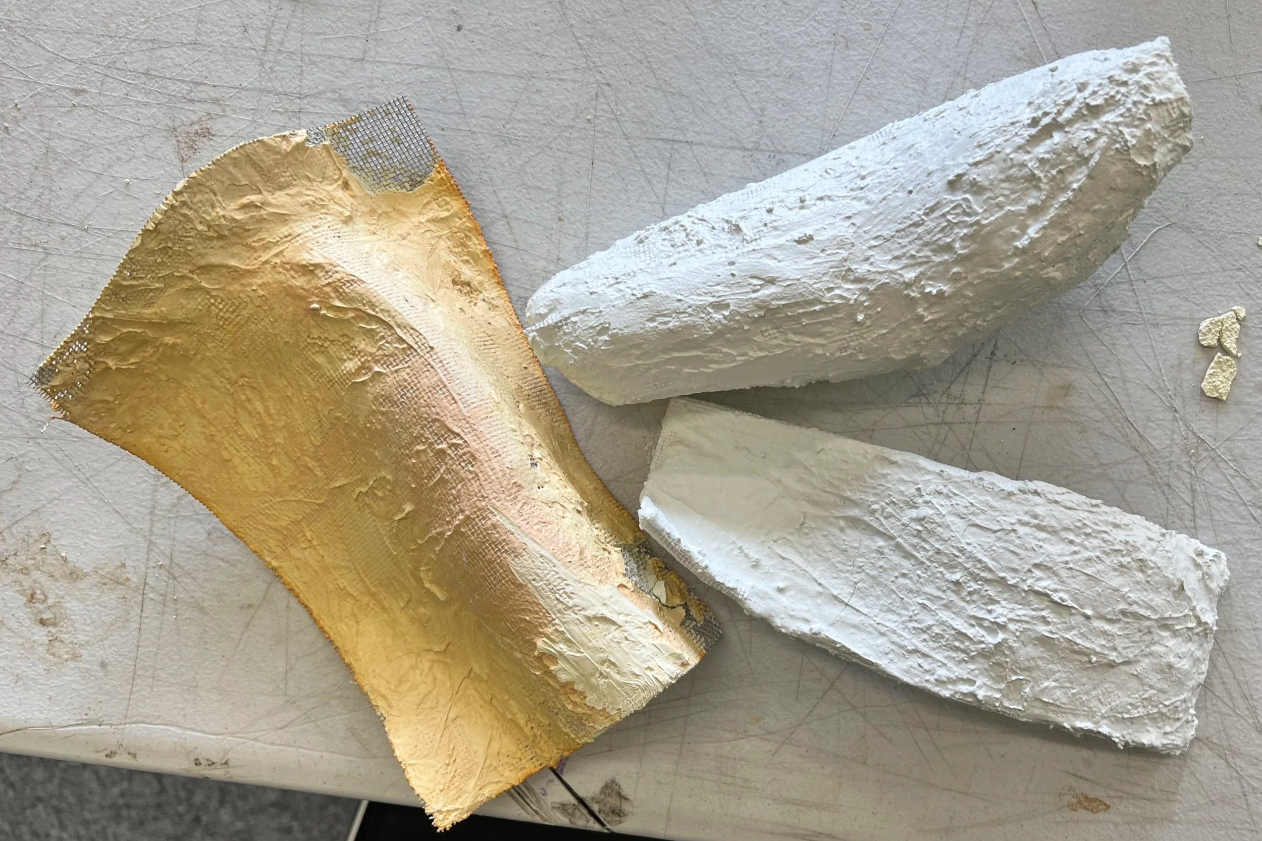 Three pieces of cheese on a white surface, including a large wedge covered with gold foil, and two blocks of white cheese with textured rinds.