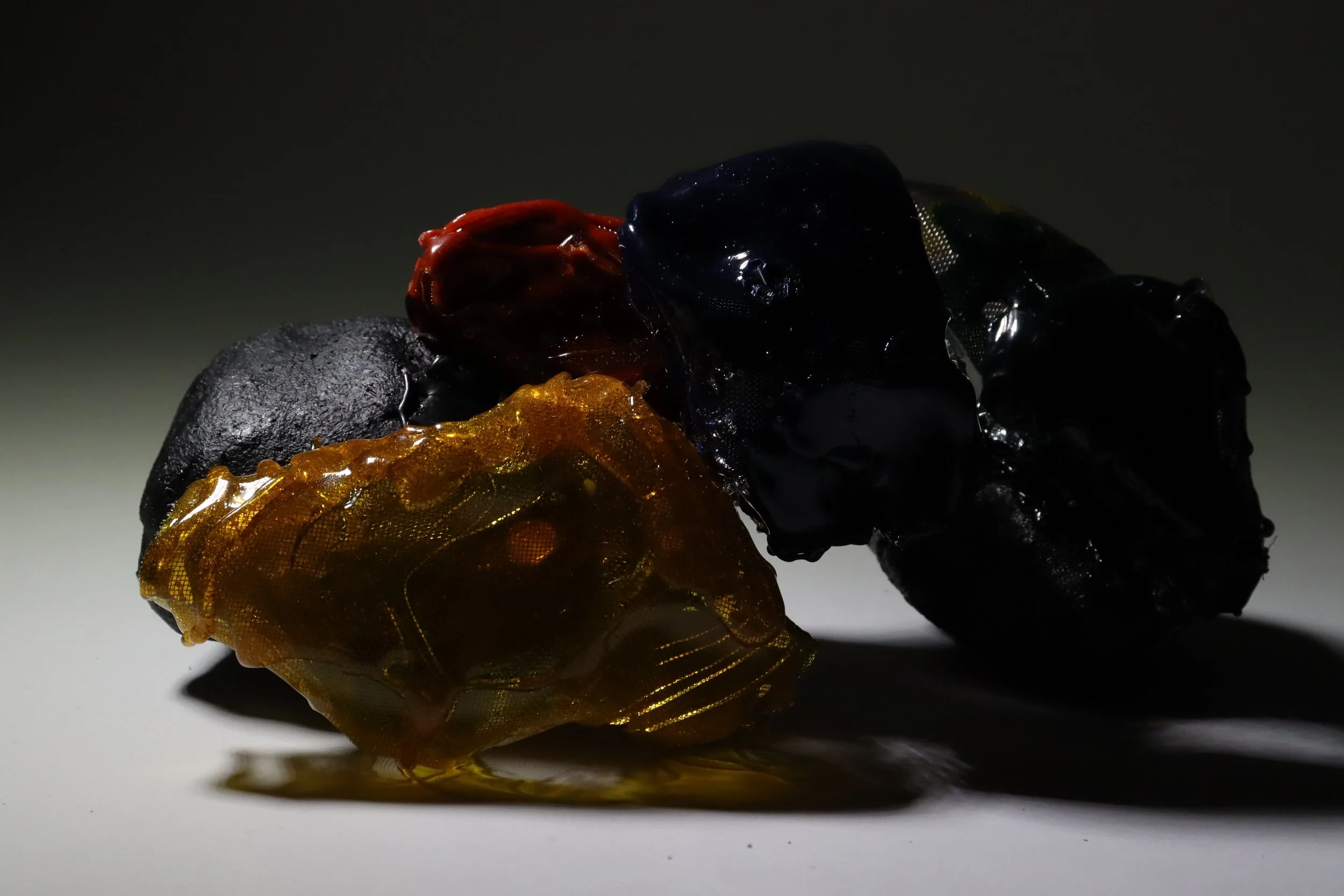 Group of three irregularly shaped, shiny, black, red, and amber colored objects on a white surface with dark background.