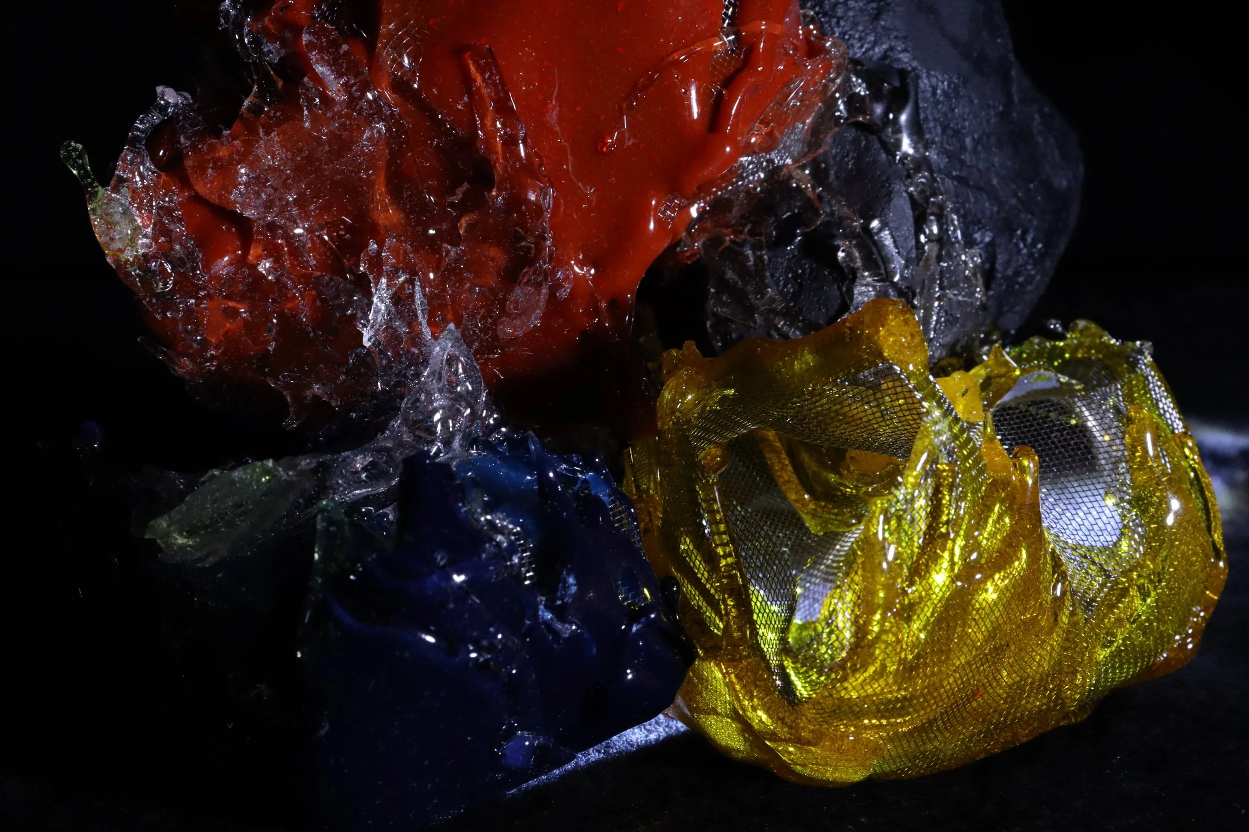 Colorful crumpled plastic or foil objects, including yellow with a mesh pattern, red, black, and blue, against a dark background.