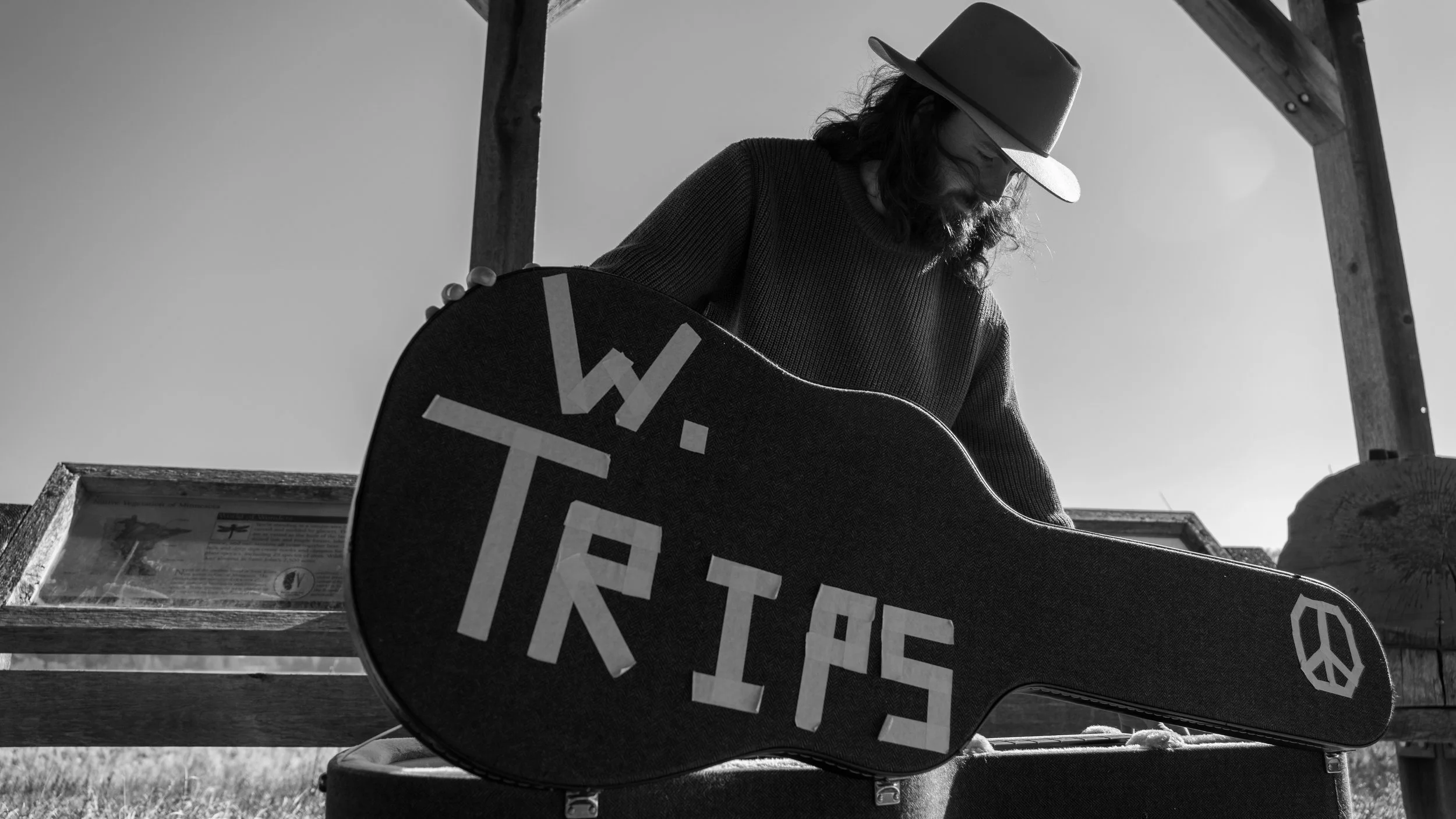 Man wearing a hat and sunglasses, holding a guitar case with 'W. TRIPS' written on it, outdoors under a wooden structure with a clear sky in the background.